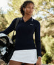 Woman wearing the Ella Knit Top in dark blue with white contrast tipping and a sleek Macade logo at the chest, styled with a white golf skort and carrying a golf bag outdoors.