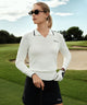 Woman wearing the Ella Knit Top in white, a long sleeve women's golf polo with a contrast-tipped open placket collar and sleek Macade logo at the chest, styled with a black skort and golf glove on a golf course. The fine knit texture and tailored fit highlight its refined, modern look for both on-course and off-course wear.