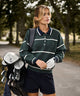 Woman wearing the Club women’s golf sweater in green with white horizontal stripes and a navy polo collar, styled with navy shorts and a golf glove while carrying a golf bag on a tree-lined course.