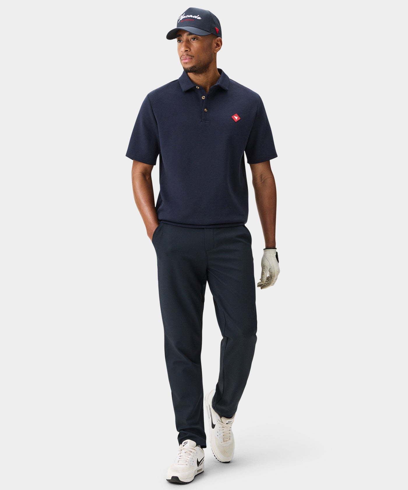 A man wears Macade Links Tech Trouser in navy with a navy polo, white sneakers, and a cap, highlighting the modern tailored fit and versatile style for golf or everyday comfort.