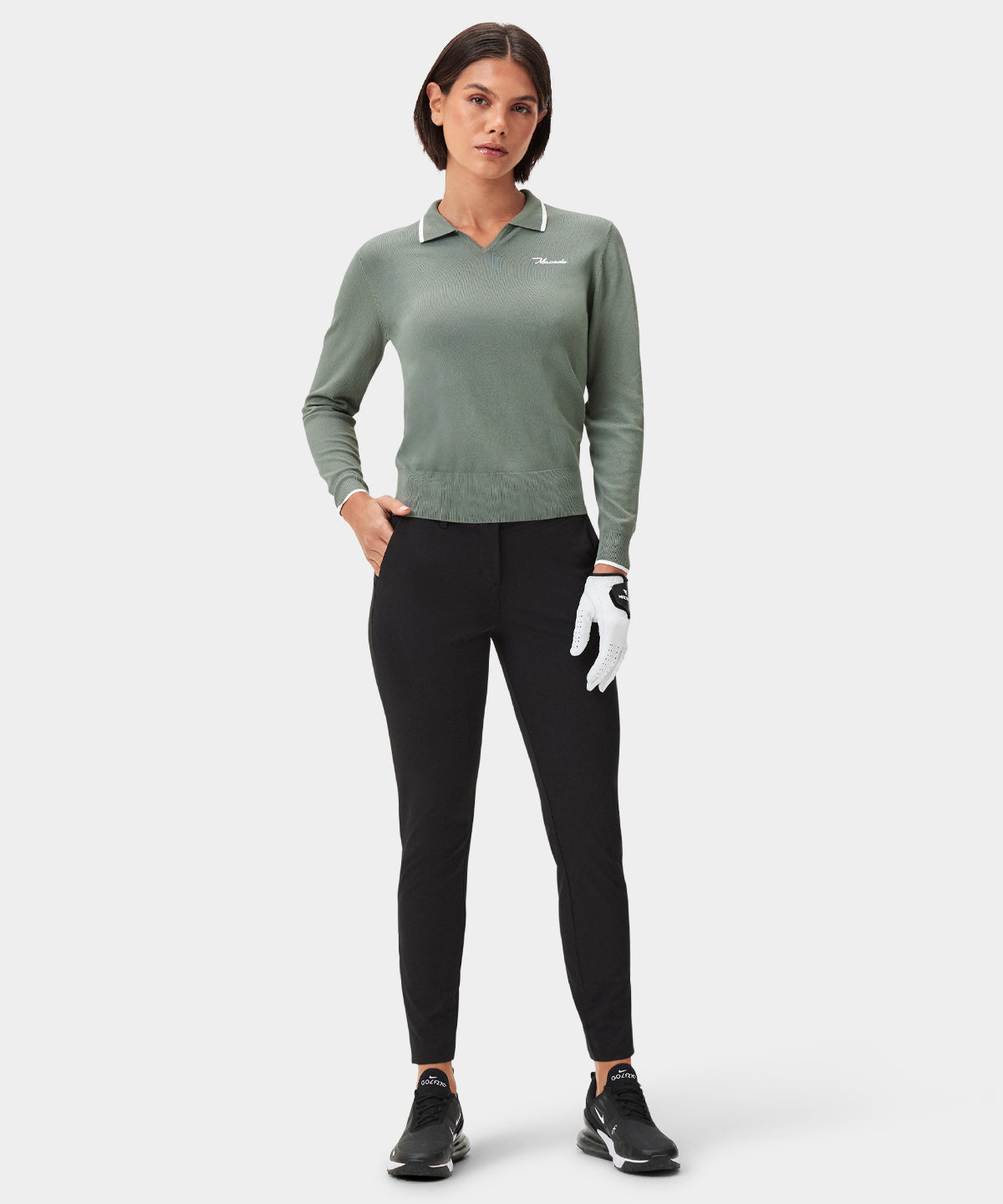 Model wearing the Ella Knit Top in sage green, featuring a long sleeve design with a contrast-tipped open placket collar, sleek Macade logo on the left chest, and ribbed cuffs, paired with black golf pants and athletic shoes for a modern, performance-driven golf outfit.
