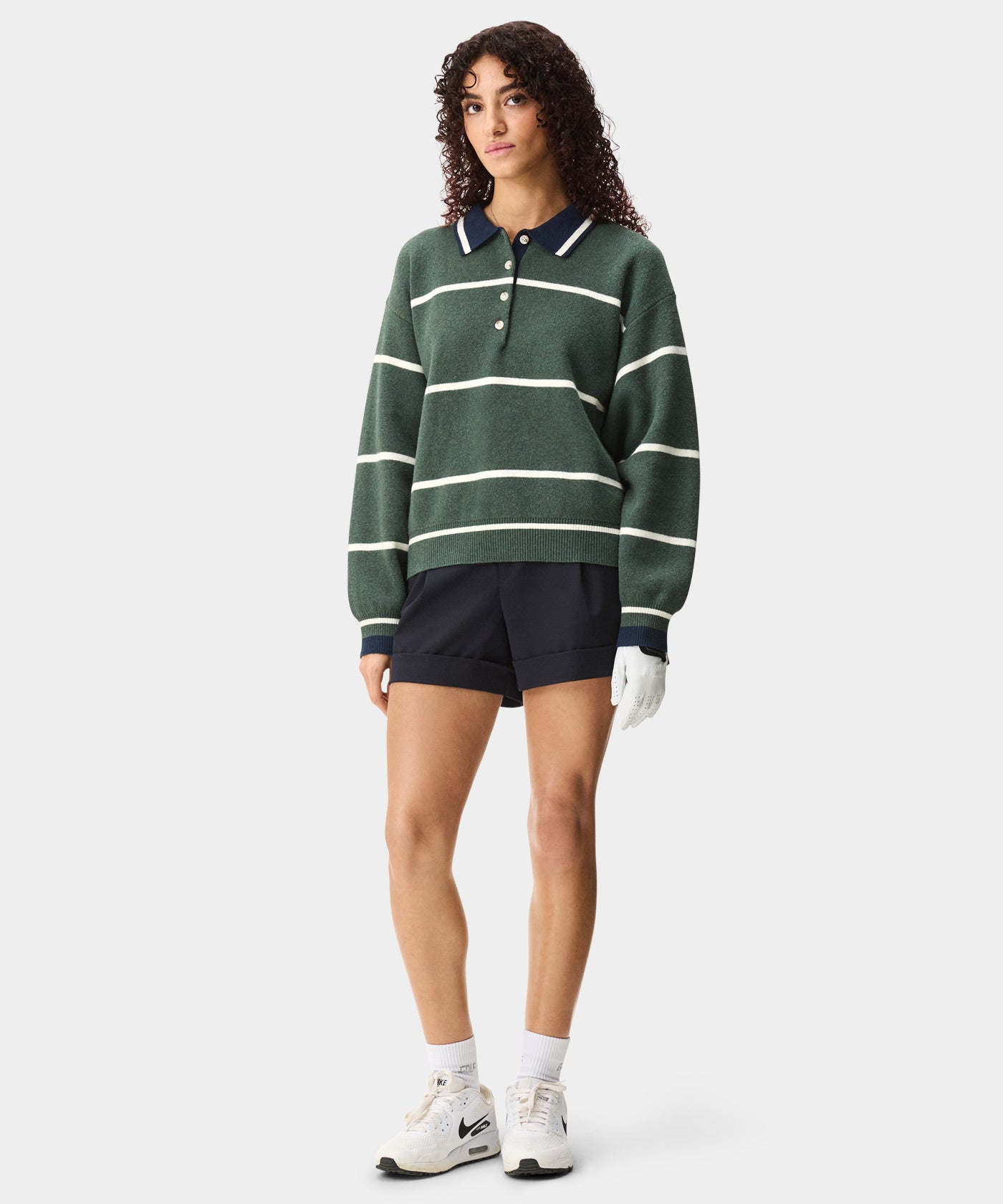 Women’s green striped golf sweater with a navy and white polo collar, button placket, and relaxed fit, styled with navy shorts, white socks, sneakers, and a golf glove for a modern, sporty look.