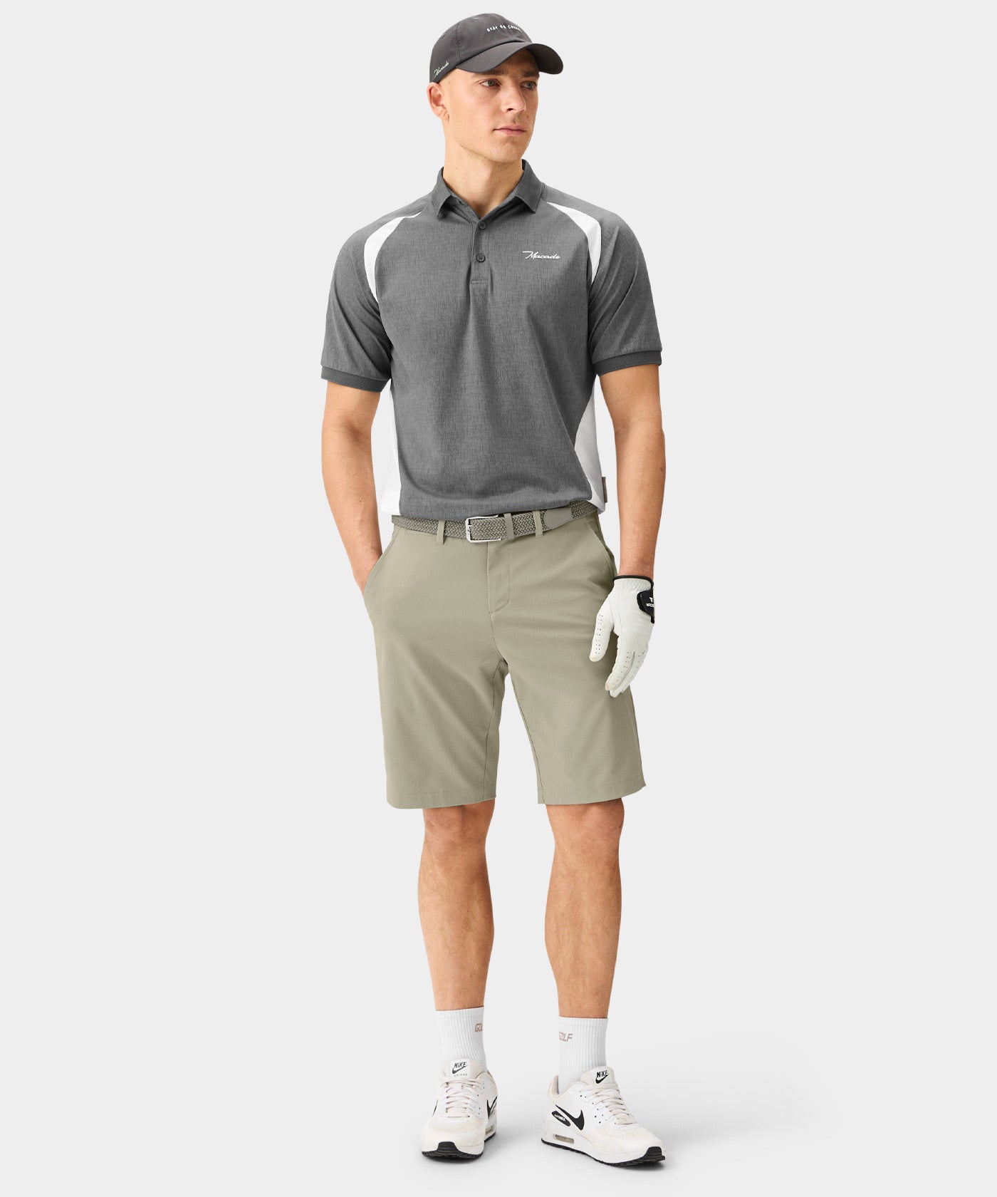A man stands wearing light olive Macade Golf Four-Way Stretch Shorts with a tailored, above-the-knee fit, styled with a grey golf polo, woven belt, golf glove, and white sneakers for a modern, athletic look on the course.