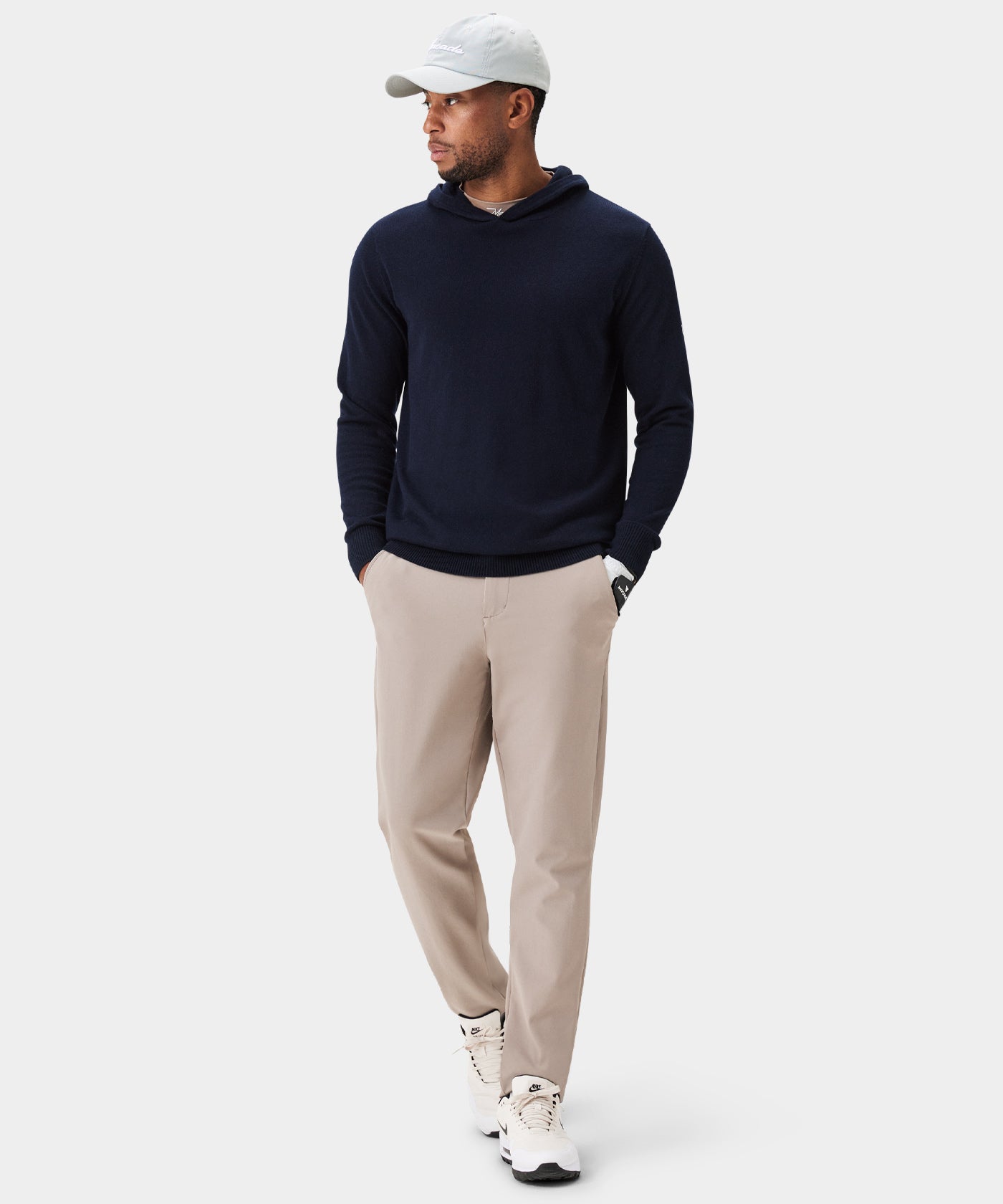 A man wears Macade Links Tech Trouser in a light tan color, styled with a navy hoodie, white sneakers, and a cap, showcasing the tailored, modern fit and clean drape of these versatile, stretch golf pants.