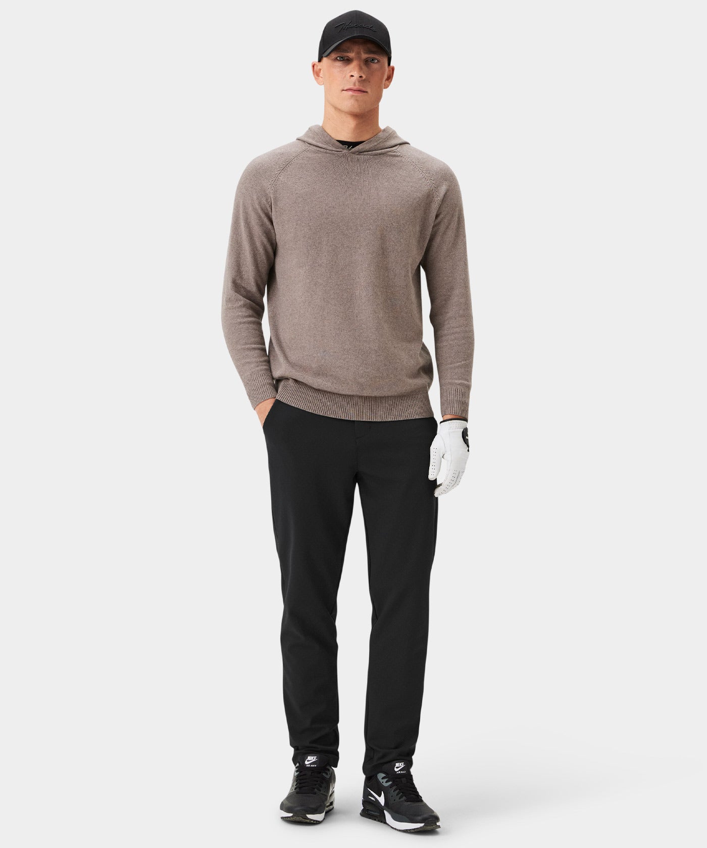 Man wearing Macade Links Tech Trouser in dark gray, styled with a taupe hoodie, black cap, and golf glove, highlighting the modern athletic fit and versatile look for both golf and casual wear.