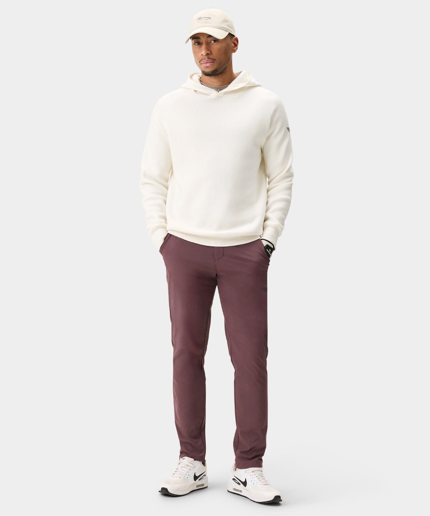 Man wearing Macade Golf Lightweight Trouser in burgundy, styled with a cream hooded sweater, white sneakers, and a beige cap, highlighting the slim, tapered fit and versatile design for both golf and casual settings.