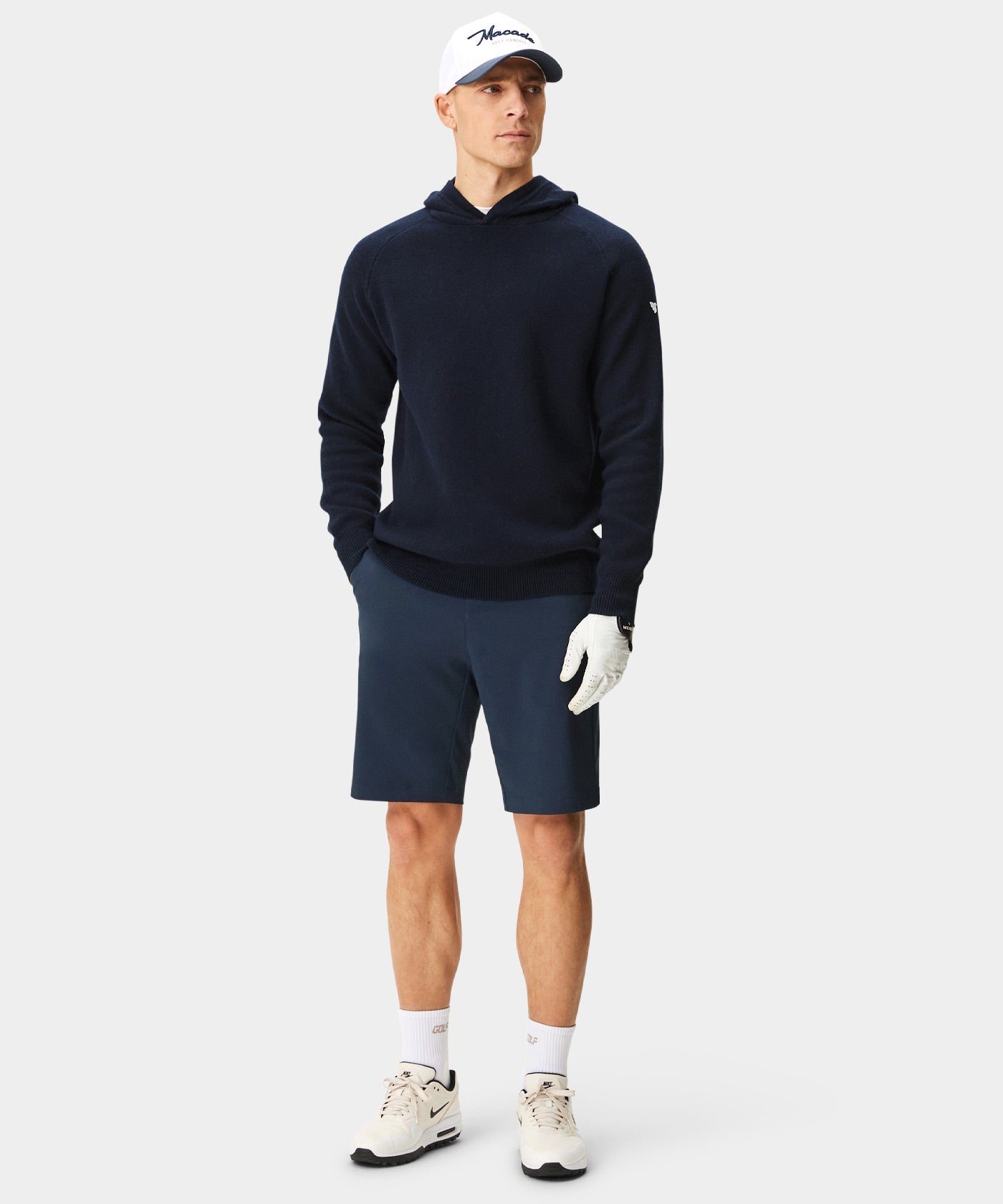 Man wearing a navy Macade Golf Tech Knit Hoodie with a modern athletic fit, paired with matching navy golf shorts, white sneakers, and a golf glove, highlighting the hoodie’s soft knit texture and minimal logo for a refined, versatile look on and off the course.