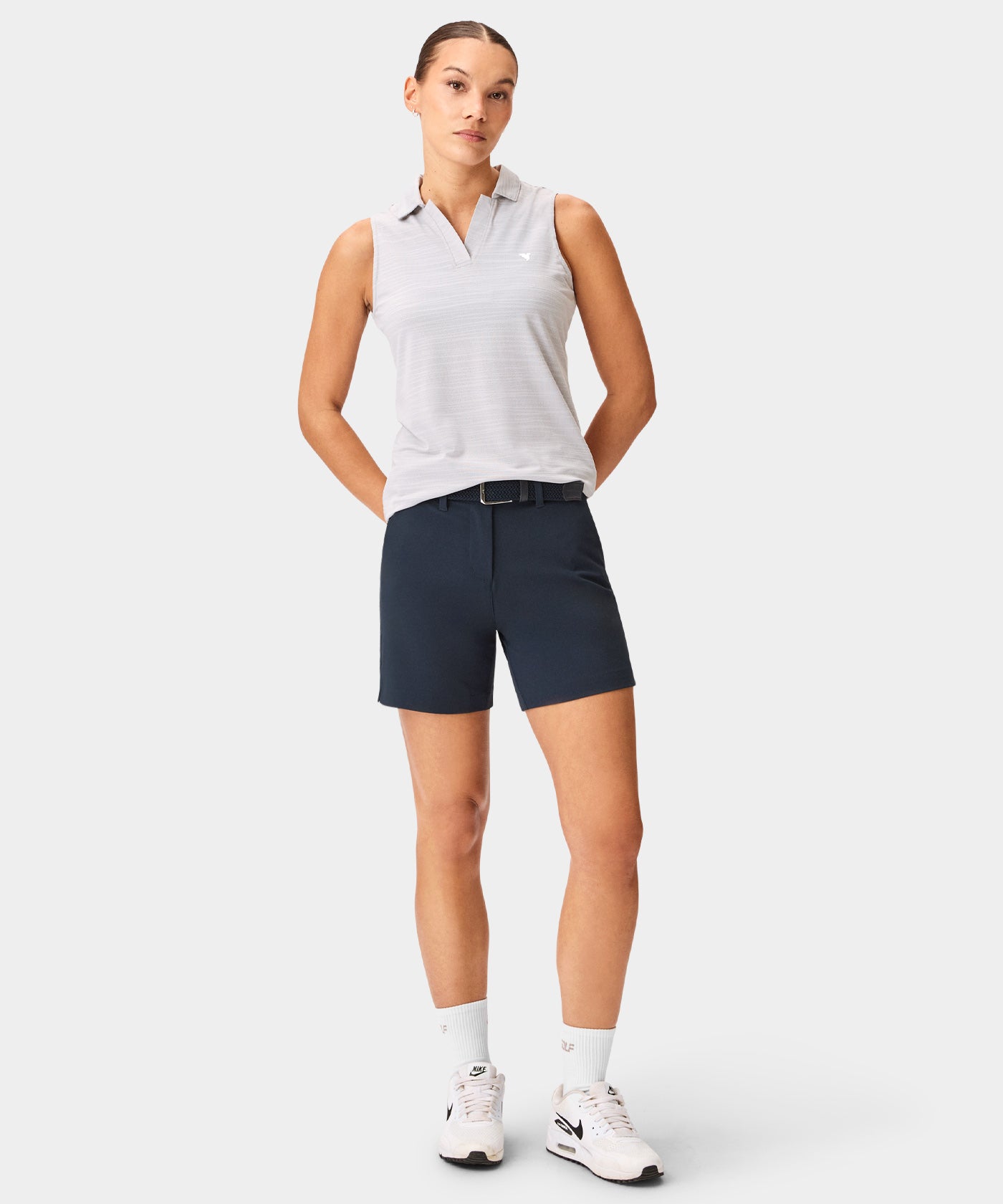 Woman wearing the Macade Paige Sleeveless Shirt in light gray, styled with navy golf shorts, white socks, and sneakers, highlighting the shirt’s clean lines, open collar, and minimal branding for a versatile, athletic golf look.