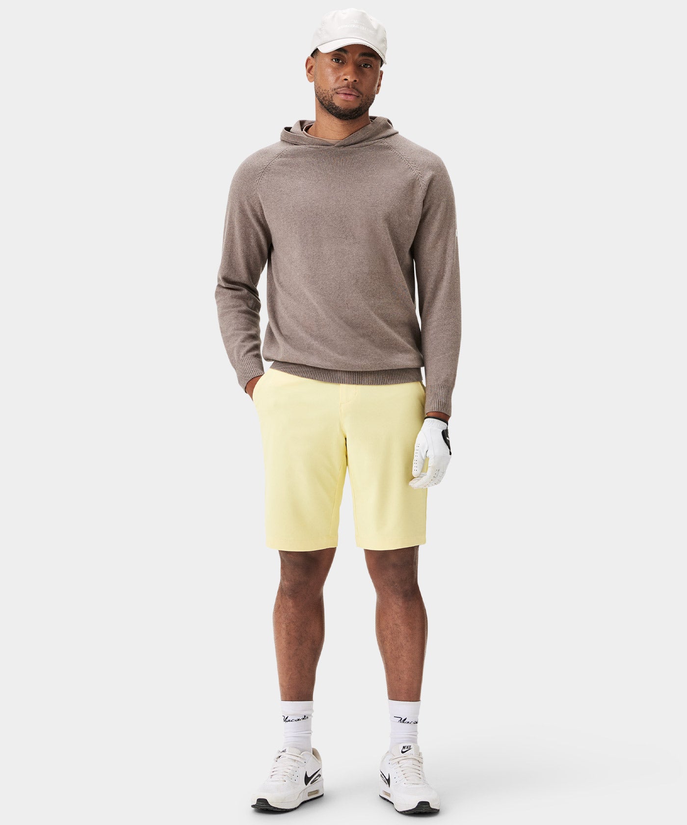 A golfer wears Macade Golf Four-Way Stretch Shorts in pale yellow, styled with a taupe hoodie, white sneakers, and golf accessories, highlighting the shorts' tailored fit, breathable fabric, and modern versatility for on and off the course.