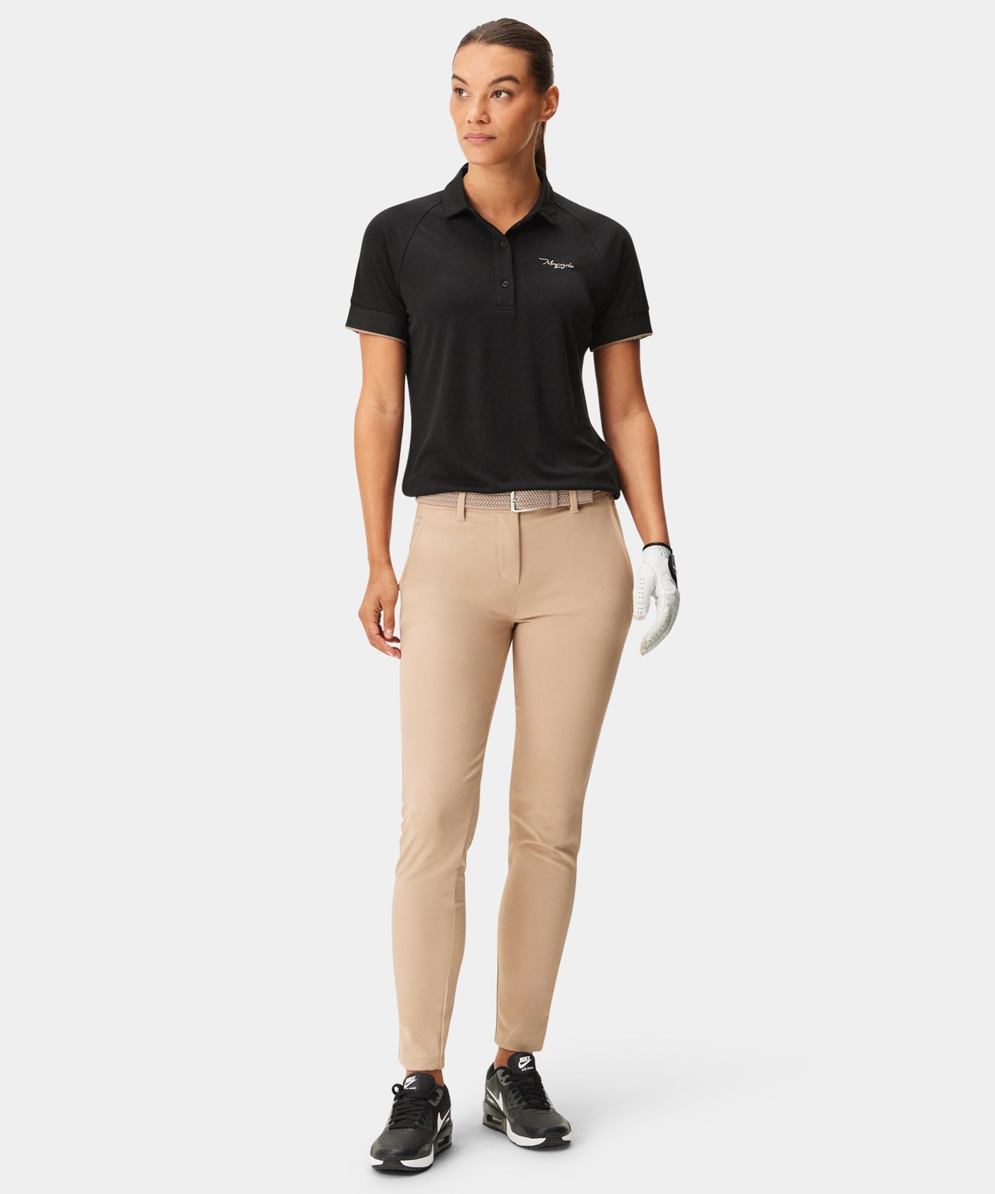 Woman wearing Macade Golf Performance Trouser in tan, styled with a black polo and golf glove, highlighting the straight, ankle-length fit, sleek belt loops, and smooth technical fabric designed for comfort and versatility on and off the course.