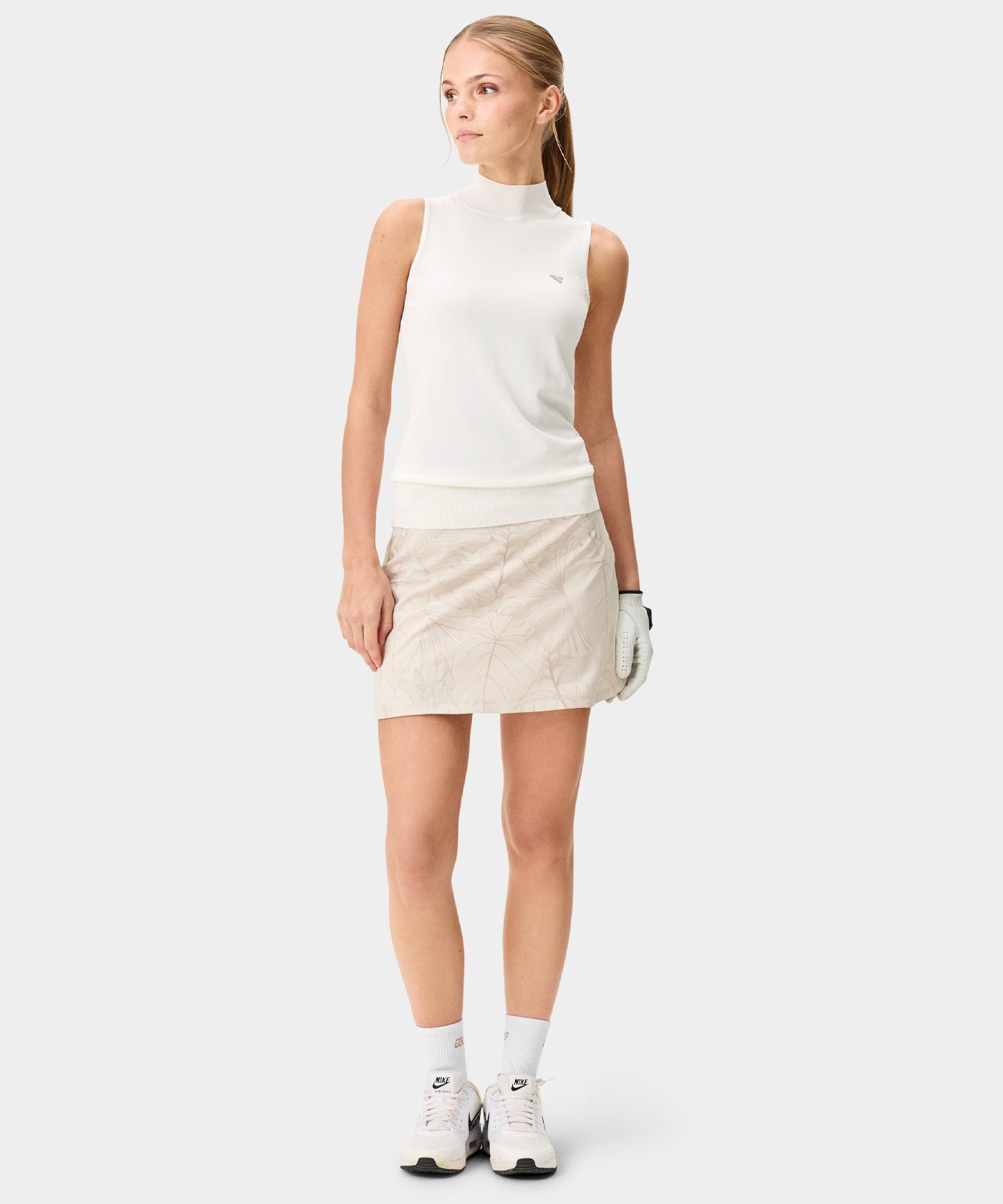 Woman dressed for golf wearing the Kaya Flex Skort by Macade Golf in a light beige leaf print, paired with a sleeveless white top, golf glove, and sneakers, showcasing the skort’s high waisted fit and minimal, athletic style.