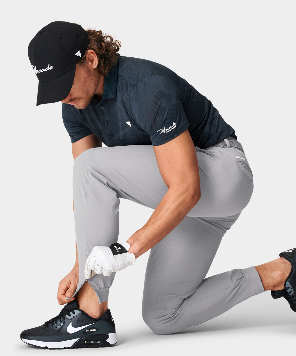 Man wearing light gray Macade Golf Four-Way Stretch Jogger, kneeling to adjust the zippered cuff at the ankle, paired with a navy golf polo and black sneakers for a modern, athletic look.