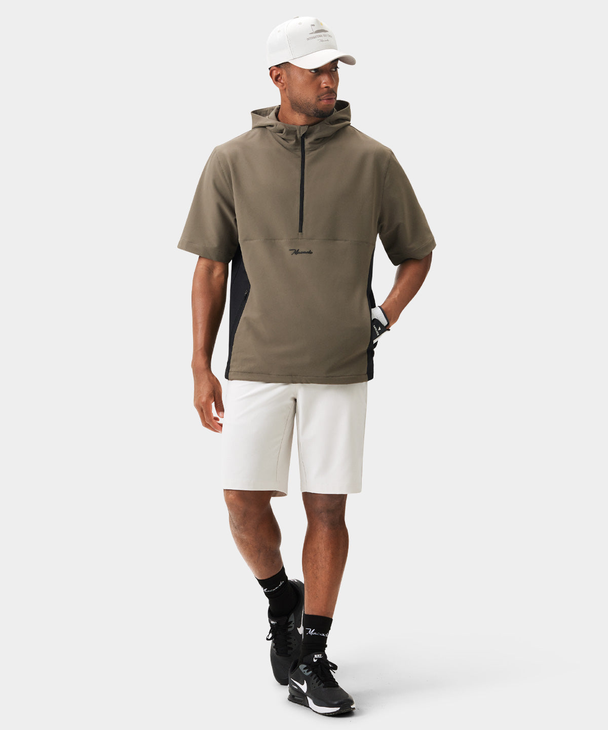 Man wearing the Macade Golf Storm Wind Shirt in olive green with a sleek half-zip front, short sleeves, and minimal branding, styled with white golf shorts, black socks, and black sneakers for a modern, athletic look.