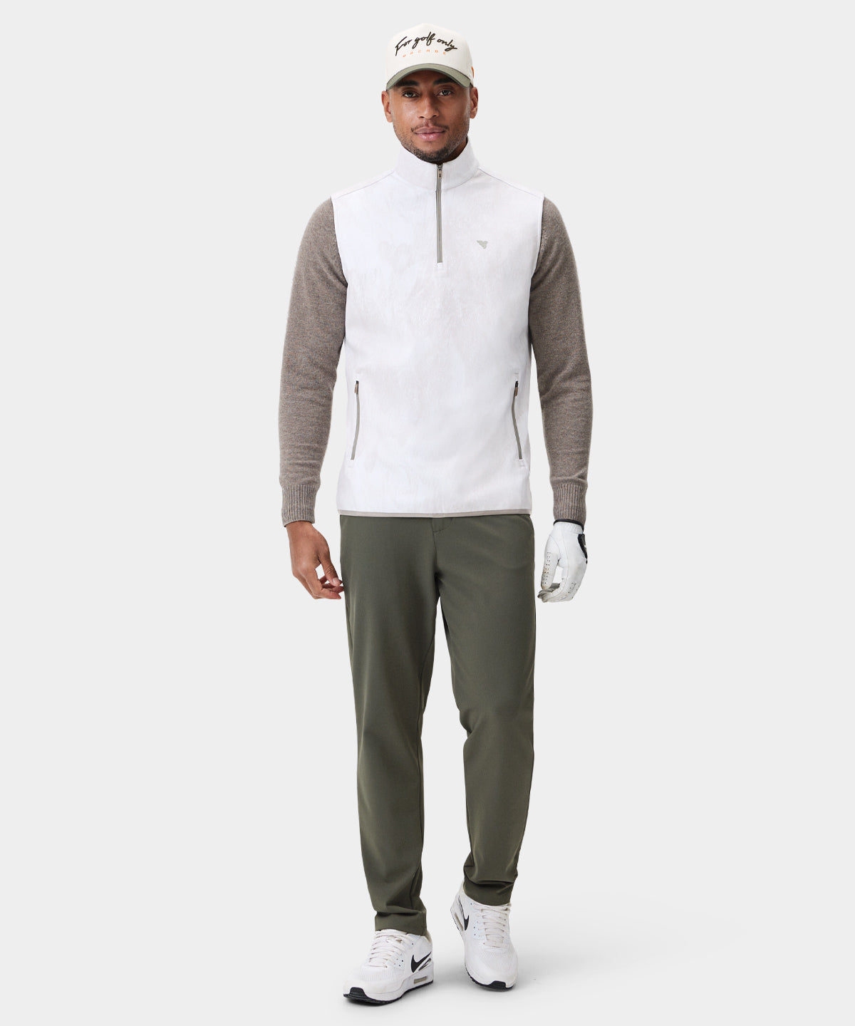 Golfer wearing the Macade Golf Therma Vest in white with a subtle pattern, featuring a high collar, quarter-zip front, and discreet zippered pockets, styled over a taupe sweater with olive green pants, white sneakers, and a golf glove for a modern, athletic look.