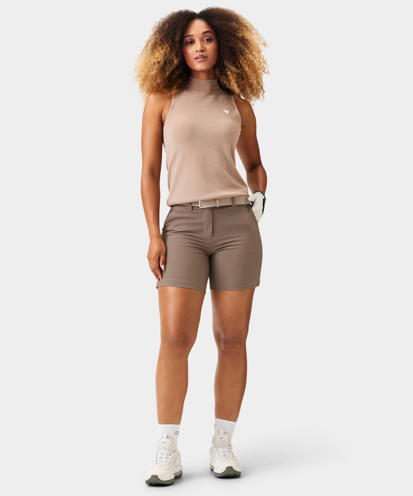 Woman wearing Macade Golf Flex Shorts in brown, styled with a beige sleeveless top, woven belt, and golf glove, highlighting the high-waisted, athletic fit and clean, tailored look designed for both golf and everyday versatility.
