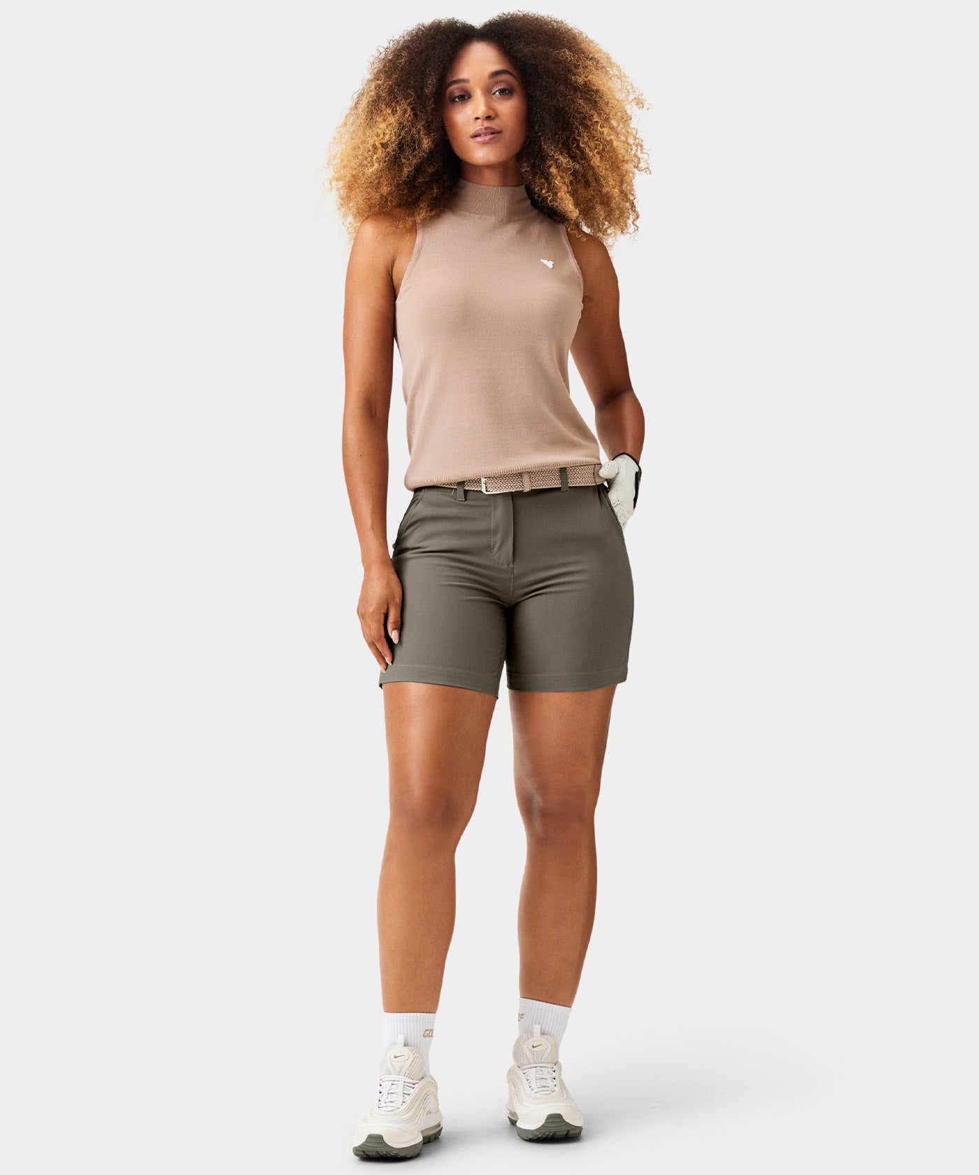 Woman wearing Macade Golf Flex Shorts in olive green, styled with a beige sleeveless top, woven belt, and athletic sneakers, highlighting the high-waisted, tailored fit and modern golf apparel look.