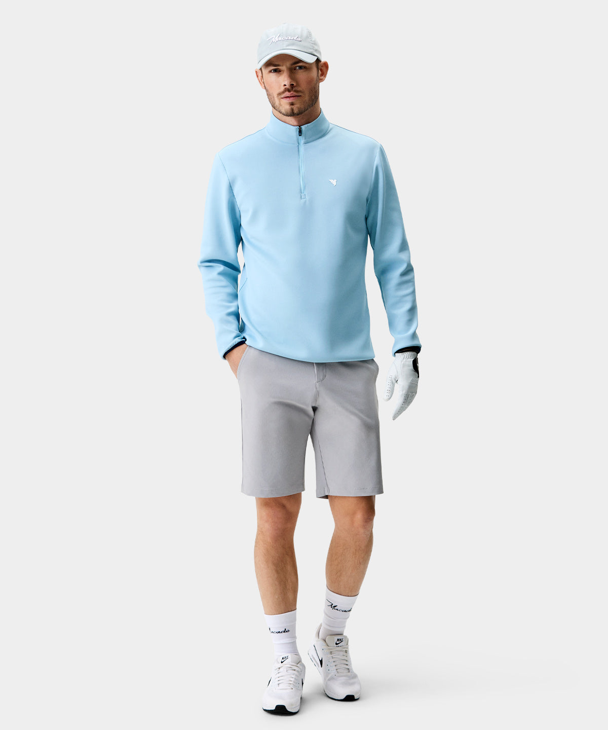 A man wears the Macade Golf Therma Quarter Zip in light blue, styled with gray golf shorts, white athletic shoes, and a golf glove, showcasing the sweater’s modern athletic fit and minimalist design for a refined look on the course.