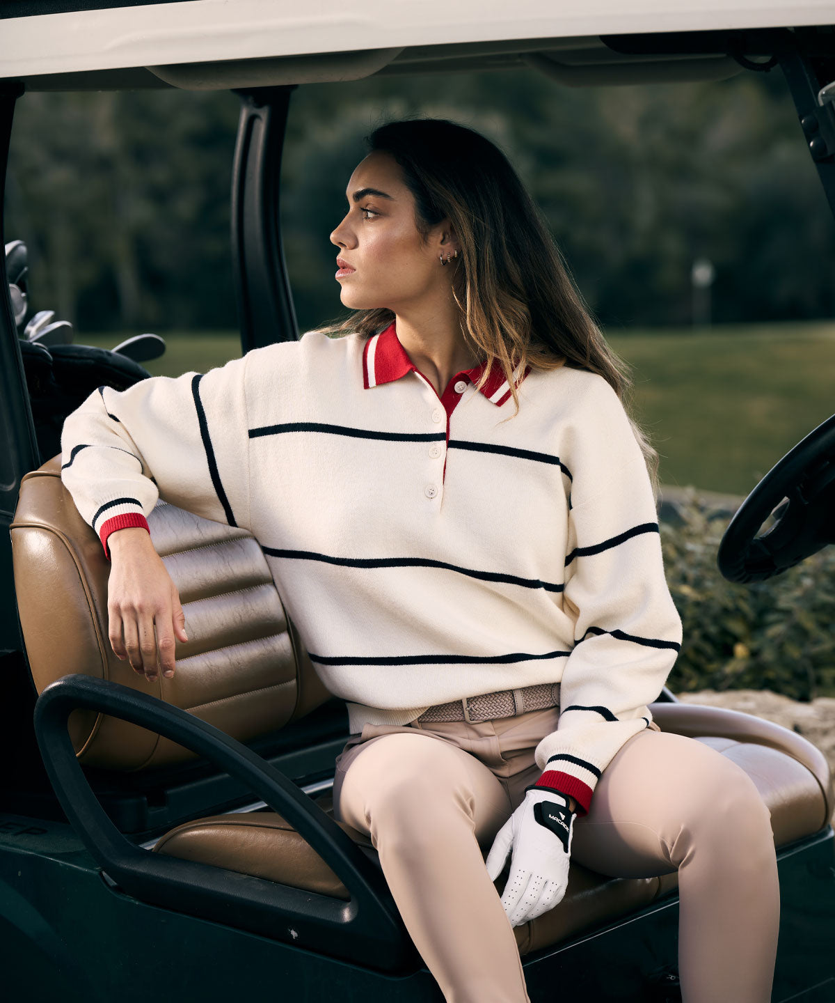 Woman sitting in a golf cart wearing the Club women’s golf sweater in ivory with navy stripes and a red polo collar, paired with beige pants and a golf glove, showcasing a refined and sporty look on the course.