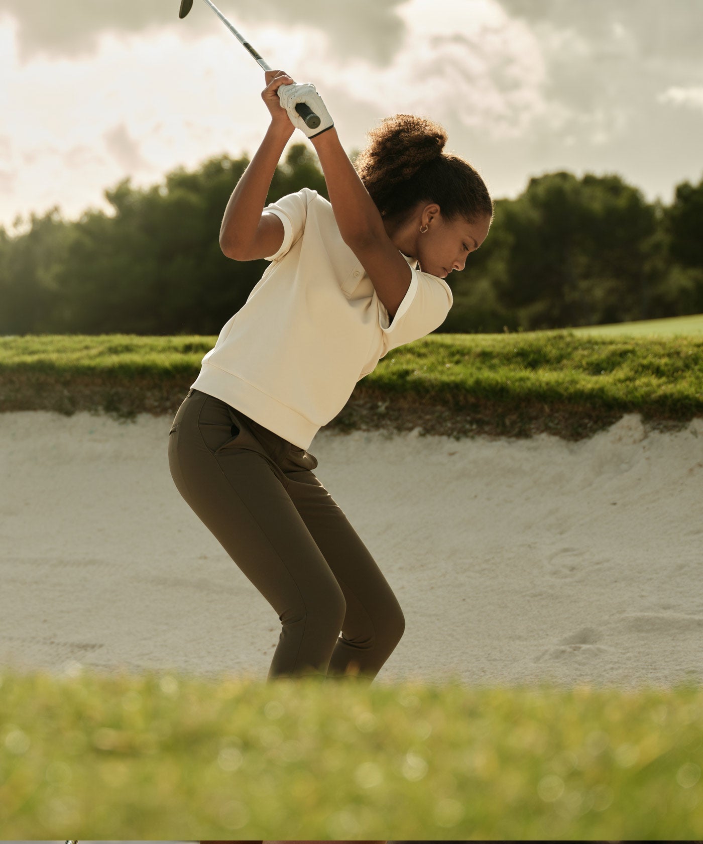 Golfer wearing the Aura TX Relaxed Shirt in tan with a white contrast collar and cuffs, swinging a club from a sand bunker, showcasing the shirt’s relaxed fit and technical stretch fabric for unrestricted motion on the course.