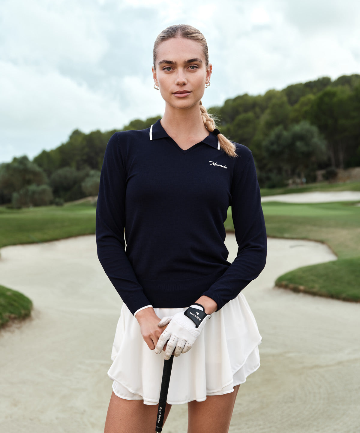 Woman on a golf course wearing the Ella Knit Top in dark blue with white contrast collar tipping and the Macade logo at the chest, paired with a white golf skort and glove for a sophisticated, athletic look.