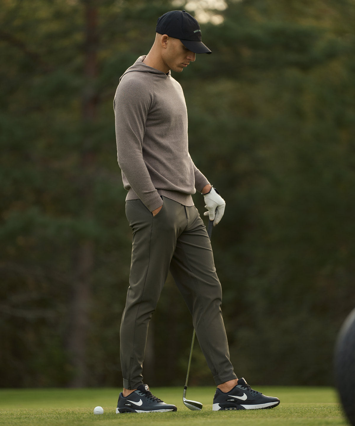 A golfer wears the Macade Golf Four-Way Stretch Jogger in olive green on the course, highlighting the athletic fit, cuffed ankles, and modern, versatile style paired with a taupe hoodie, golf glove, and sneakers for a seamless transition from play to everyday life.
