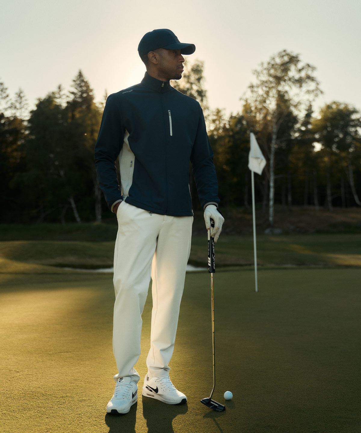 Golfer on the green wearing the Macade Golf Storm Wind Jacket in dark blue with light gray side panels, paired with white pants and golf shoes, highlighting the jacket’s tailored fit and modern, performance-focused design.