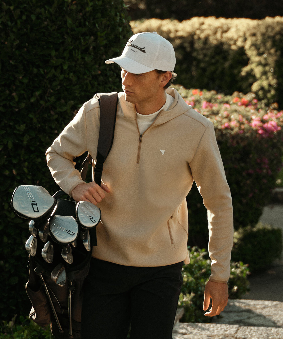 Man wearing the Therma Zip Hoodie in Tan, carrying a golf bag outdoors, highlighting its athletic fit, high neckline, and minimalist design for versatile comfort on and off the course.