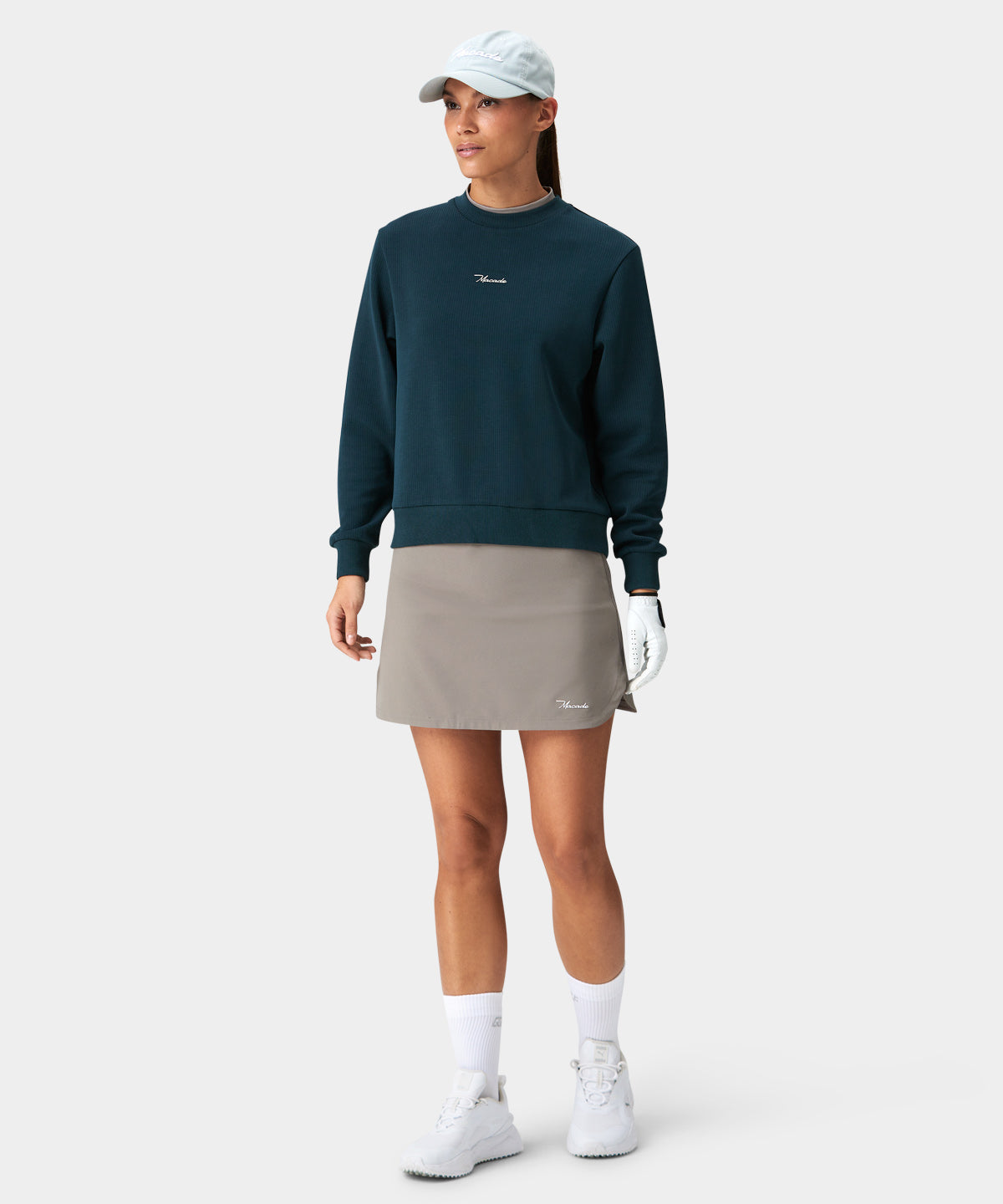 Woman wearing the Kaya Flex Skort by Macade Golf in taupe, styled with a dark teal sweatshirt, white golf shoes, and a cap for a versatile, modern golf outfit.