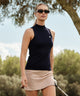 Woman wearing a navy Kayla Air Sleeveless Top with a high mock neck and ribbed hem, paired with a tan golf skirt, standing outdoors on a golf course holding a club.