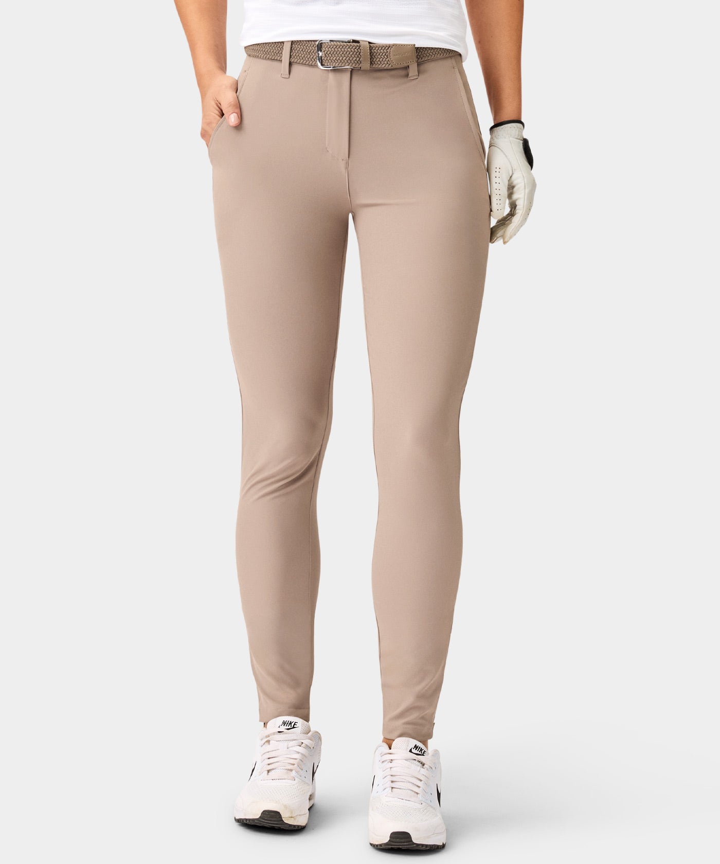 A woman models Macade Golf Four-Way Stretch Joggers in brown, featuring a tailored athletic fit, ankle-length cut with subtle cuffs, deep pockets, and a textured belt, styled with white sneakers and a golf glove for a clean, modern look.