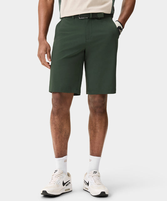 A man wears dark green Macade Golf Four-Way Stretch Shorts with a tailored, above-the-knee fit, styled with a woven belt, golf glove, and white sneakers for a refined, athletic look on and off the course.