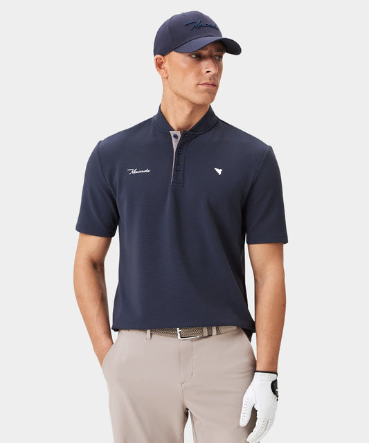 Man wearing a deep navy Macade Golf Heath Bomber Shirt with a bomber-style collar, minimal white branding, and a contrasting button placket, paired with light beige golf pants, a matching belt, a navy cap, and a golf glove for a refined athletic look.