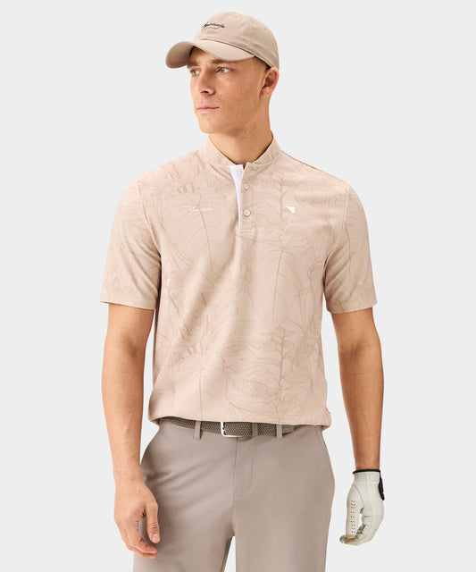 Man wearing the Macade Golf Heath Bomber Shirt in tan with a subtle palm print, featuring a bomber-style collar, minimal white branding, and short sleeves, paired with taupe golf trousers, a matching cap, and a golf glove for a refined, modern golf look.