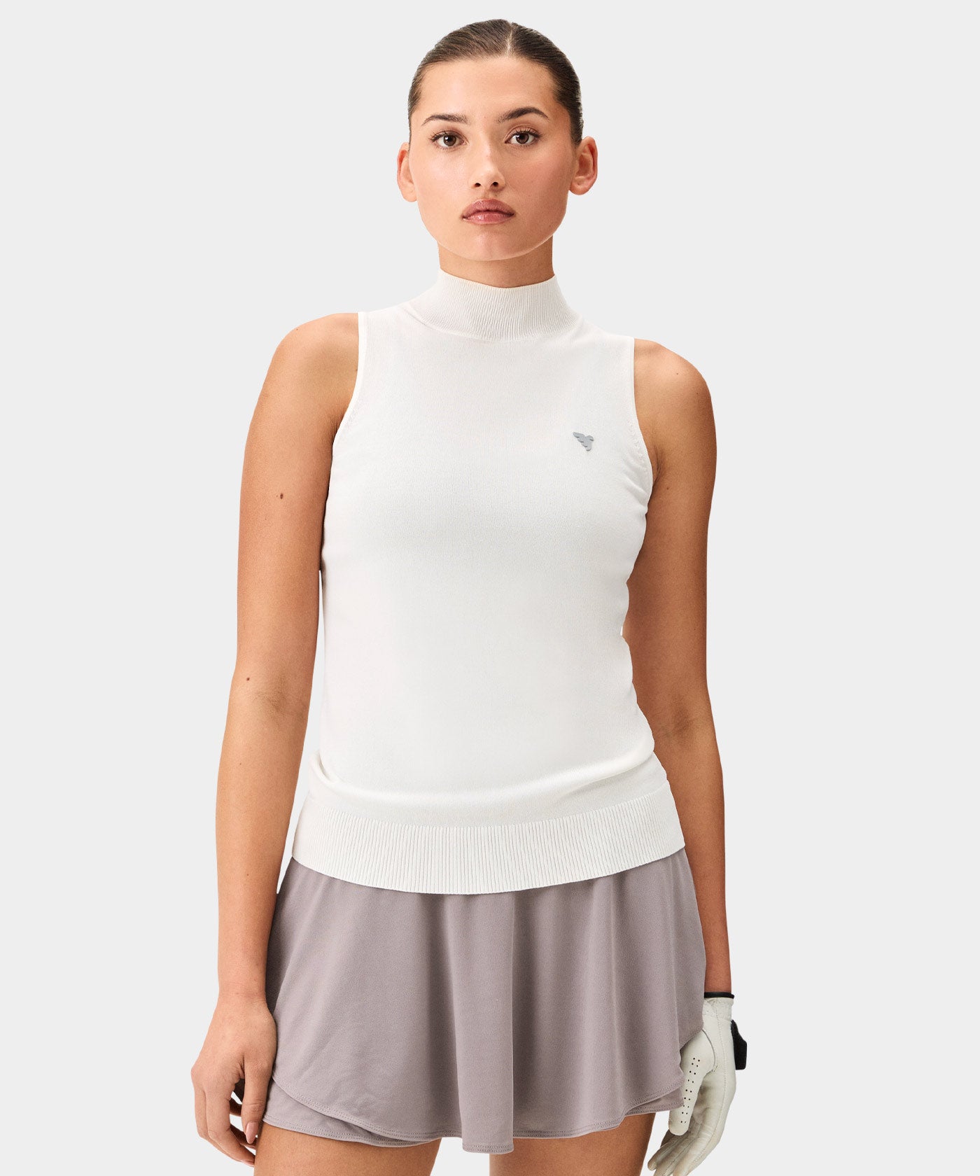 Woman wearing a white Kayla Air Sleeveless Top with a high mock neck, ribbed hem, and subtle chest logo, paired with a light taupe golf skirt and a white golf glove for a clean, modern golf look.