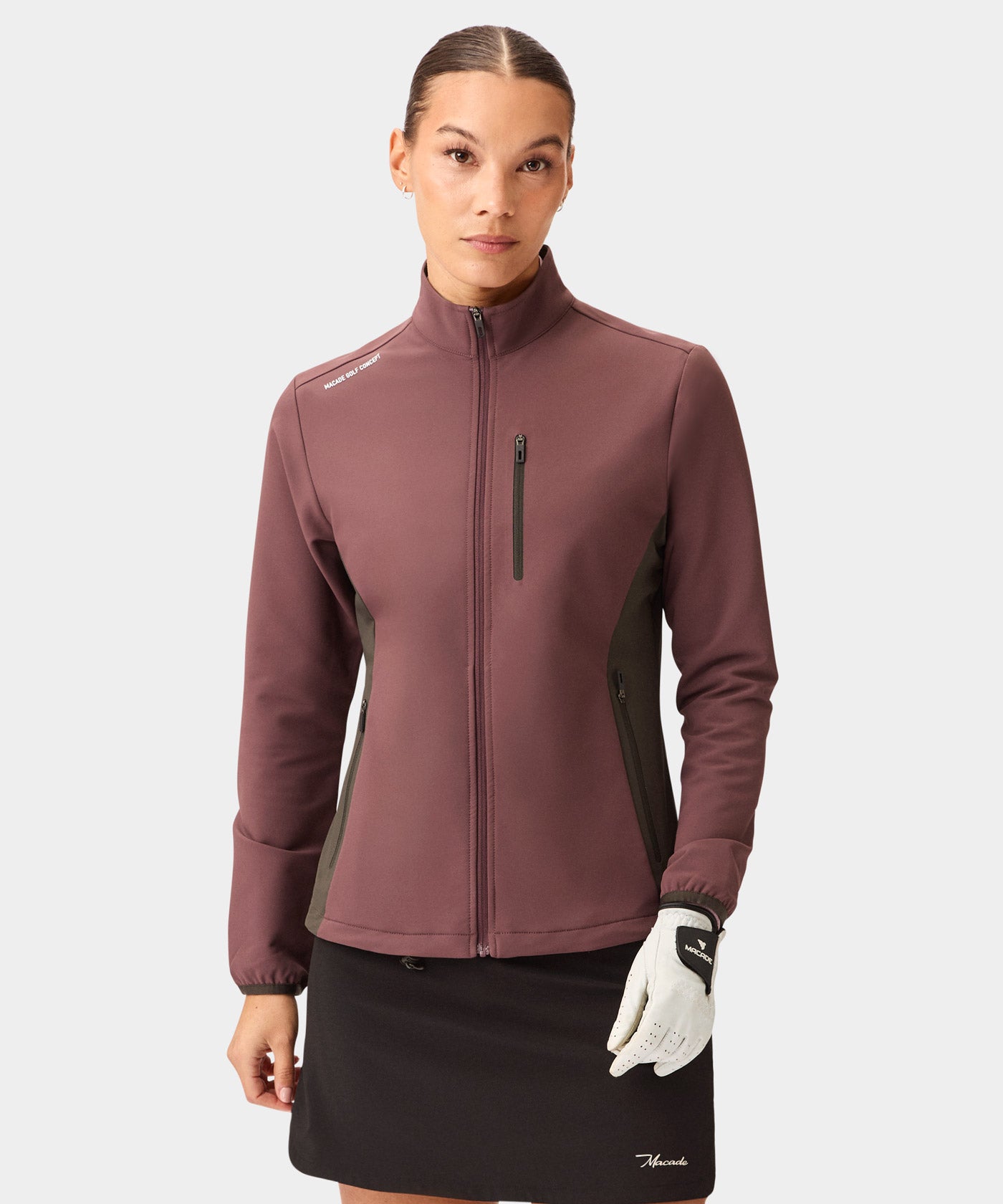 A woman models the Macade Golf Storm Wind Jacket in mauve with dark olive side panels, featuring a full zip front, high collar, and sleek chest pocket, styled with a black golf skirt and glove for a modern, athletic look.