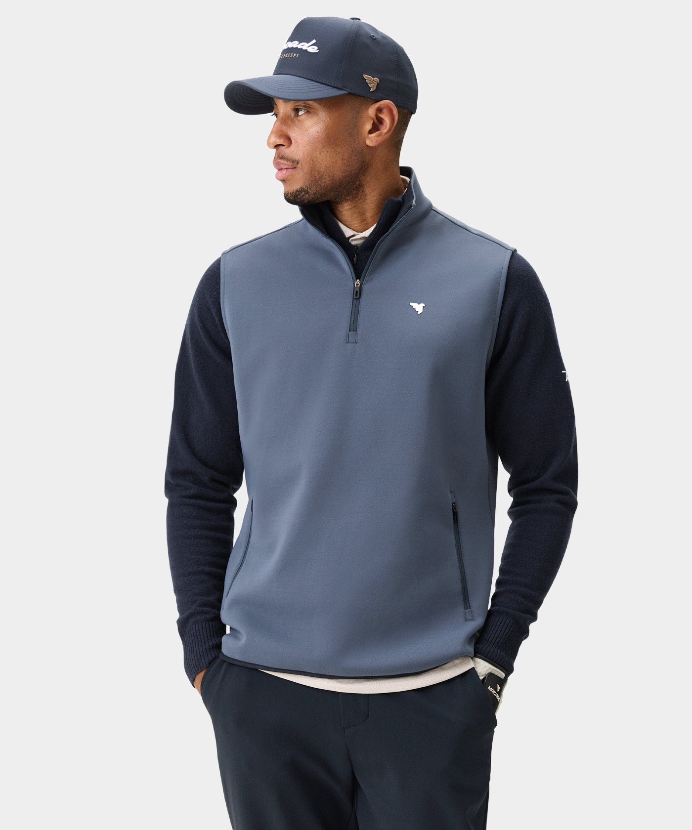 Man wearing the Macade Golf Therma Vest in Stone Blue, layered over a navy sweater with matching navy pants and a Macade cap, highlighting the vest’s modern fit, high collar, and discreet zip pockets for a polished golf look.