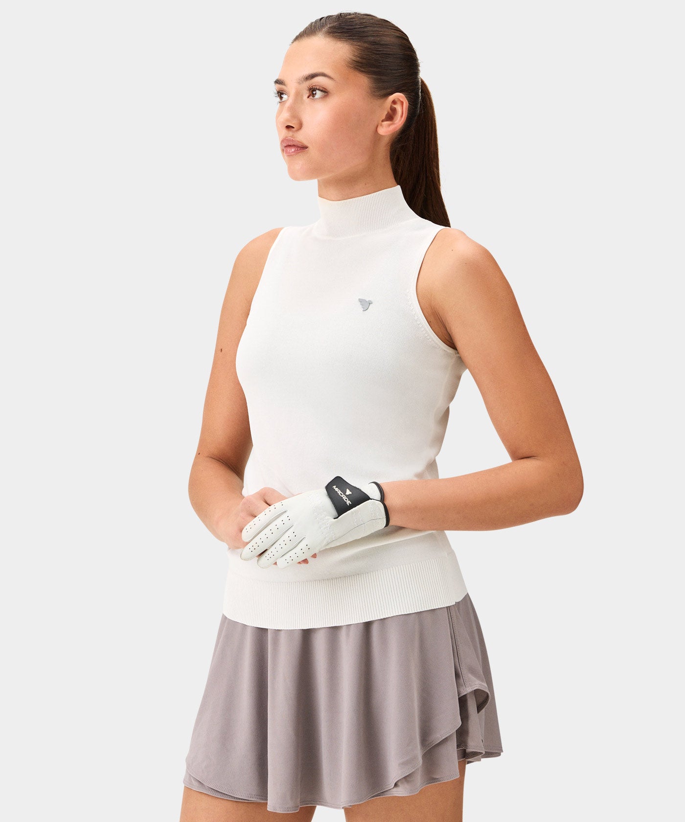 Woman in a white Kayla Air Sleeveless Top with a high mock neck and ribbed hem, paired with a taupe golf skirt and golf glove, highlighting the top’s soft knit texture, minimal branding, and modern fit for a refined golf or resort look.
