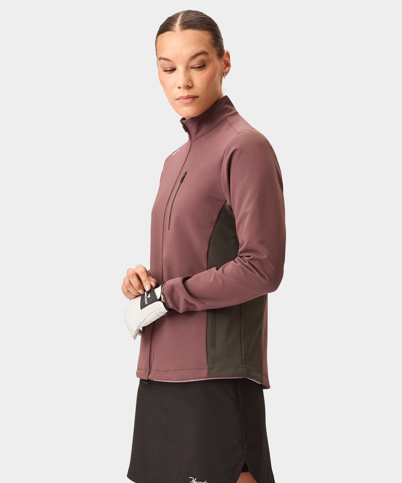 A woman wears the Macade Golf Storm Wind Jacket in mauve with dark olive side panels, featuring a full zip front, high collar, and zippered chest pocket, styled with a black golf skirt and glove for a modern, athletic look.