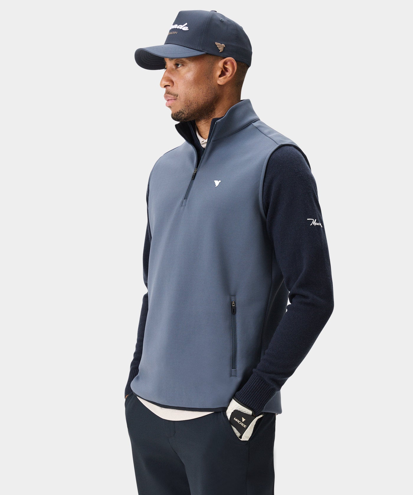 A man models the Stone Blue Macade Golf Therma Vest layered over a navy sweater, paired with dark pants and a matching cap, highlighting the vest’s modern fit, high collar, and minimalist design for versatile golf and everyday wear.