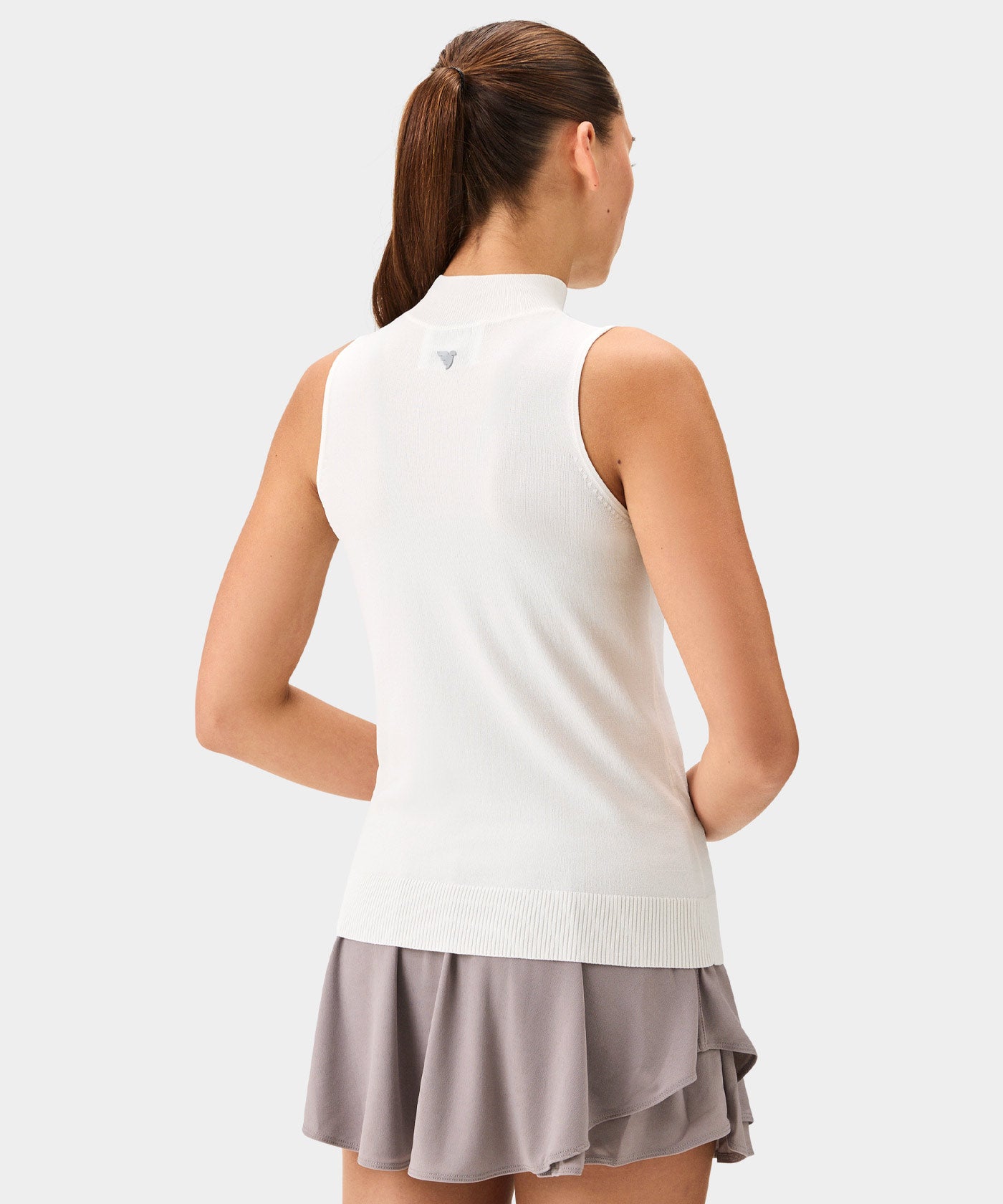 Back view of a woman wearing a white Kayla Air Sleeveless Top with a high mock neck, ribbed hem, and minimal branding at the upper back, paired with a light taupe golf skirt for a clean, modern golf style.