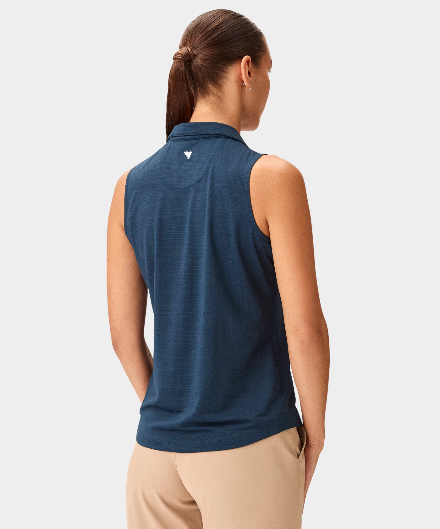 Back view of a woman wearing the Macade Paige Sleeveless Shirt in steel blue, featuring a clean, minimal design and lightweight fabric, paired with beige golf pants for a refined and versatile golf outfit.