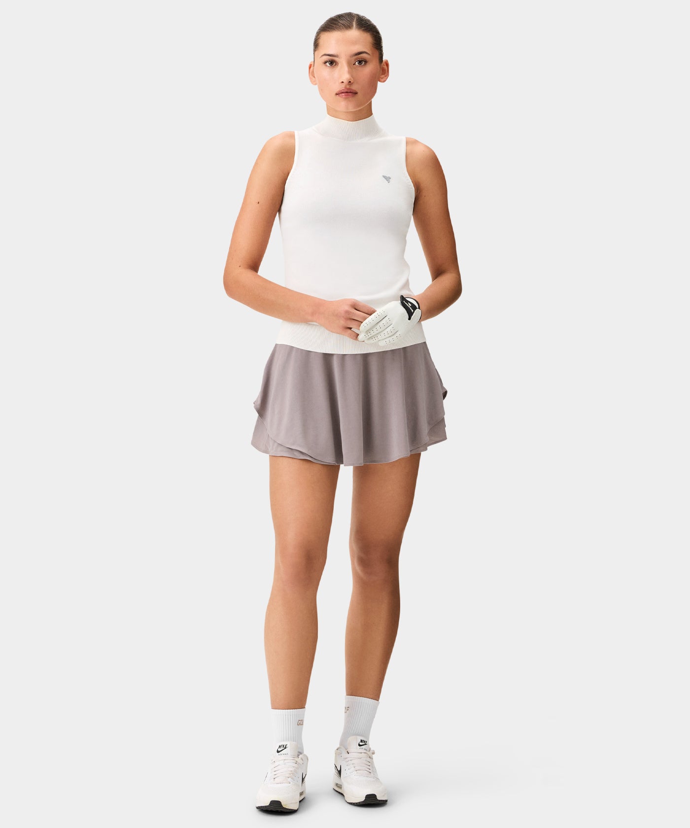 Woman wearing a white Kayla Air Sleeveless Top with a high mock neck and ribbed hem, paired with a flowy taupe golf skirt, white socks, and sneakers, highlighting the top’s clean silhouette, minimal branding, and lightweight knit for a fresh, versatile golf look.
