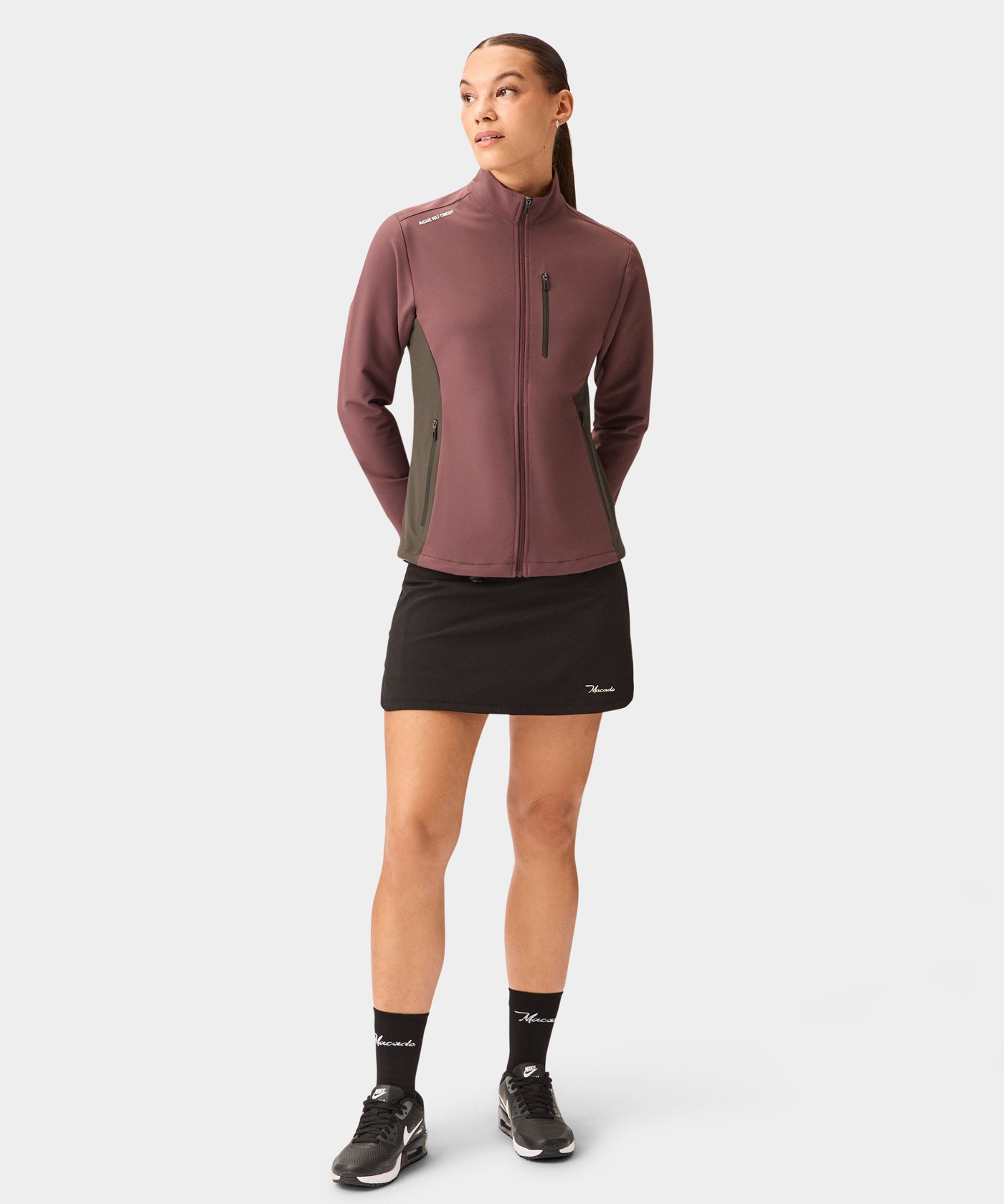 A woman models the Macade Golf Storm Wind Jacket in mauve with dark contrast side panels, featuring a tailored fit, zippered chest and side pockets, and sleek minimal branding, paired with a black golf skirt and athletic sneakers against a plain background.