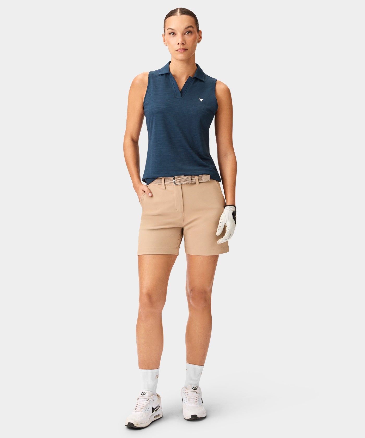 Woman wearing the Macade Paige Sleeveless Shirt in steel blue, styled with beige golf shorts, white socks, sneakers, and a golf glove, highlighting the shirt’s clean lines, open collar, and minimal branding for a modern, athletic look.