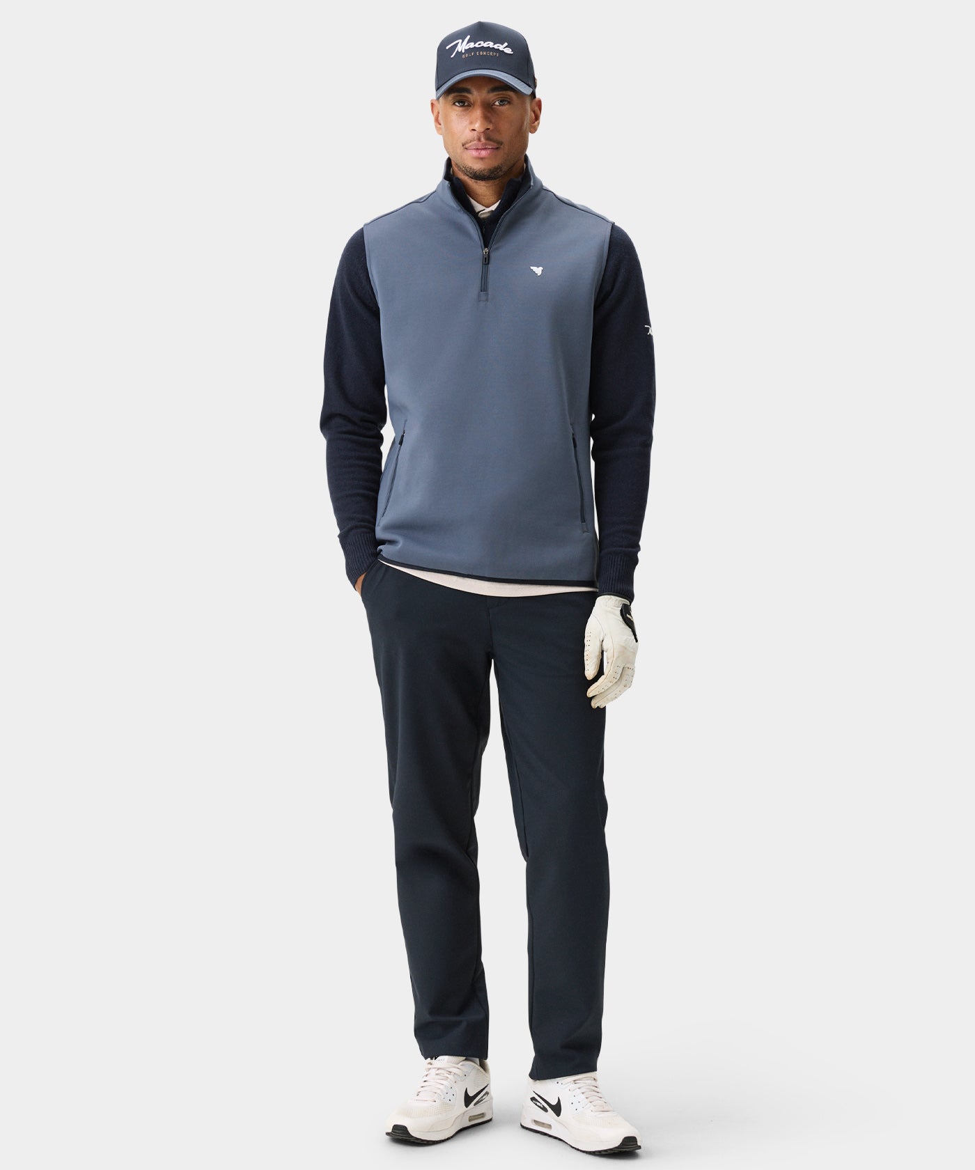 Male golfer wearing the Macade Golf Therma Vest in Stone Blue layered over a navy sweater, styled with navy pants, white sneakers, a Macade cap, and a golf glove for a modern, versatile look on and off the course.