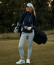 A woman golfer carries a golf bag while wearing the Macade Golf Storm Wind Jacket in dark blue with off-white contrast side panels, styled with light golf pants, a cap, and golf glove, highlighting the jacket’s tailored fit, zippered pockets, and sleek minimal branding on the course.